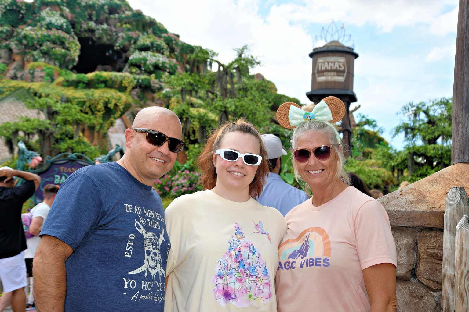 Three family members smiling near Tiana's Bayou Adventure at Magic Kingdom, with the attraction water tower in the background